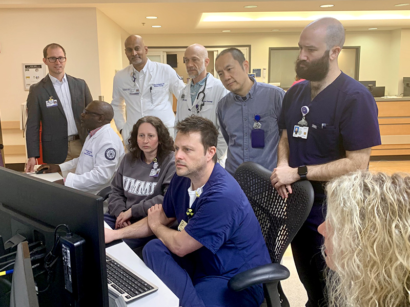 A group of healthcare professionals gather around a computer workstation in a clinical area, reviewing information on a screen.